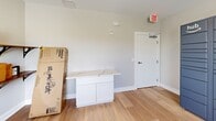 Located at 105 Pomeroy Ave in Meriden, Connecticut, this dedicated package room serves as a centralized mail and delivery management hub for residents of Pomeroy Apartments. The room is finished with light hardwood flooring and neutral-toned walls, creating a clean and well-maintained environment. A standout feature is the Amazon Hub smart locker system, which lines one wall and provides residents with secure, self-service package retrieval around the clock. Opposite the lockers, a set of wall-mounted wooden shelves with black metal brackets offers organized storage for additional parcels and deliveries of varying sizes. A built-in white cabinetry unit with a stone or quartz countertop provides a practical surface for sorting and processing packages. Natural light enters through a window fitted with blinds, supplementing the recessed ceiling lighting. The room is equipped with a ceiling vent for climate control, a smoke or security sensor, and a clearly marked exit door with a door closer for safety compliance. The overall layout is efficient and functional, designed to streamline the package receiving process for building residents.