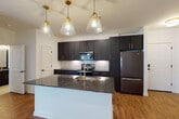 This single-floor residence showcases a contemporary design with a cohesive blend of dark, high-contrast finishes and clean white walls throughout. The kitchen serves as a central focal point, featuring floor-to-ceiling dark navy shaker-style cabinetry accented with gold hardware, dark granite countertops, and a herringbone-patterned tile backsplash. A generously sized center island with an undermount sink provides additional workspace and casual seating. Stainless steel appliances, including a refrigerator, range, and microwave, complete the kitchen's modern aesthetic. A row of clear glass pendant lights with gold fixtures hangs above the island, adding a refined decorative element. The kitchen opens seamlessly into the living area, which benefits from natural light streaming through glass double doors leading to an outdoor balcony or patio. Rich wood-look flooring runs throughout the main living areas, while bedrooms feature plush neutral carpet and ceiling fans. Additional rooms include a well-appointed bathroom with dark vanity cabinetry, granite countertops, light tile flooring, and a glass-enclosed walk-in shower with matte black fixtures. The overall ambiance is modern and move-in ready, with thoughtful finishes throughout.