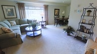 This single-floor space showcases an open-concept layout that seamlessly connects the living and dining areas, creating a cohesive and inviting environment. The living area is anchored by plush, wall-to-wall carpeting in a soft neutral tone, which adds warmth and comfort underfoot. A large picture window dressed with elegant draping curtains floods the space with abundant natural light, enhancing the airy feel of the room. The sofa seating is positioned to take advantage of the natural light, accompanied by a stylish round coffee table with a lower display shelf, adding both function and visual interest.

Transitioning naturally into the dining area, a dark-finished dining table paired with matching chairs provides a defined space for meals and gatherings. A framed artwork piece on the wall and a tasteful floral arrangement on the table contribute to the polished, welcoming atmosphere. The neutral wall tones and consistent flooring throughout unify the two areas, while decorative plants placed strategically around the space bring touches of natural greenery. Overall, the space exudes a calm, well-maintained ambiance suited for comfortable everyday living.
