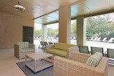 This covered community lounge is a thoughtfully designed outdoor gathering space that bridges indoor comfort with open-air living. The area features textured stucco walls and a smooth covered ceiling fitted with a ceiling fan, keeping the space comfortable in warm weather. The lounge is furnished with wicker-style seating, including a sofa and armchair with green and striped cushions, arranged around a low coffee table to create an inviting conversation area. Additional folding chairs provide flexible seating for residents and guests. Large open-air openings on multiple sides allow for generous natural light and views of the surrounding landscaped grounds, including mature trees and a decorative iron railing that borders an adjacent patio area. A sparkling pool is visible just beyond the covered structure, reinforcing the amenity-rich atmosphere of the community. Dark-painted doors provide access to utility or restroom facilities nearby. A dedicated alcove houses a drinking fountain alongside safety signage, a first aid kit, and an emergency information board, reflecting a well-maintained and safety-conscious environment. A no-smoking sign is posted prominently, indicating a clean and considerate shared space. Overall, this lounge area offers a welcoming, functional retreat for residents to unwind and socialize outdoors.