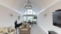 This refined single-floor space is defined by its most striking architectural feature: a soaring barrel-vaulted ceiling with decorative arched molding that lends the interior a grand, cathedral-like quality. A statement chandelier with candle-style lighting hangs at the center of the vault, complemented by wall sconces that provide warm, ambient illumination throughout.

The living area is thoughtfully arranged with a plush, neutral-toned sofa and armchair accented by patterned throw pillows, centered around a round glass-top coffee table atop a soft area rug. A wall-mounted flat-screen television sits above a dark wood media console, flanked by sconces for a balanced, polished look. Framed botanical artwork adds a touch of nature-inspired elegance to the walls.

Adjacent to the living area, the dining space features a round pedestal table with dark wood cross-back chairs, positioned beneath the spectacular arched window that floods the room with natural light and offers views of the surrounding greenery. Decorative plate wall art and potted plants add warmth and character to the space.

A set of white-framed French doors opens to an adjoining area, enhancing the sense of openness and connectivity. Neutral gray carpeting and soft wall tones tie the entire space together into a cohesive, inviting, and sophisticated environment.
