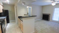 This single-floor residence offers an open, connected layout that integrates the kitchen and living areas into one cohesive space. The kitchen is anchored by a peninsula with dark countertops and light wood cabinetry, providing both workspace and a natural dividing point between the kitchen and adjoining areas. A track lighting fixture overhead illuminates the kitchen, ensuring well-lit food preparation and casual dining. The flooring transitions from tile in the kitchen zone to carpeting in the living area, subtly defining each space while maintaining visual continuity. The living area is generously proportioned and features a fireplace with a tiled surround, serving as a natural focal point for the room. Neutral-toned walls throughout contribute to a clean, understated ambiance that complements a variety of décor styles. A doorway visible from the kitchen provides access to additional areas of the unit, while a pass-through window connects the kitchen to a secondary space, enhancing the overall sense of openness. The layout is practical and efficient, making good use of the available square footage.