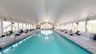 Located in Liberty Lake, Washington, this single-story indoor pool facility offers a resort-style aquatic experience in a bright, well-appointed setting. The centerpiece is a generously sized lap pool with crystal-clear turquoise water, framed by a wide concrete pool deck on both sides. Rows of rattan-style lounge chairs line the perimeter, providing ample space for relaxation before or after a swim. The vaulted ceiling rises overhead, enhancing the sense of openness and airiness throughout the space. Abundant natural light pours in through large windows that run along multiple walls, offering panoramic views of the surrounding landscape, including outdoor courts and manicured grounds. A dedicated lounge area features wicker seating with side tables, ideal for casual gatherings or quiet relaxation. At one end of the pool, a spa or hot tub is visible, adding another layer of wellness amenity. The entry area includes glass doors, a recycling station, and posted facility rules, reflecting a well-managed community environment. Ceiling-mounted speakers and wall-mounted monitors suggest the space is equipped for entertainment. The overall ambiance is clean, modern, and inviting, with a neutral color palette and thoughtful design that creates a tranquil indoor retreat.