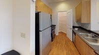 Located at 13200 York Rd in Charlotte, NC, this single-floor unit offers a practical and well-organized layout with a variety of functional living spaces. The kitchen is equipped with stainless steel appliances, including a refrigerator, range, and dishwasher, complemented by warm wood-toned cabinetry, dark countertops, and durable hardwood-style flooring. A door from the kitchen leads to an adjoining utility or pantry area, adding convenient storage. The living areas transition to neutral-toned carpet, which extends throughout the bedrooms. Each bedroom features a ceiling fan with light fixture and windows dressed with horizontal blinds, allowing natural light while maintaining privacy. One bedroom benefits from a tray ceiling detail, adding a subtle architectural element. The bathroom includes a soaking tub with a tiled shower surround, hardwood-style flooring, and ample cabinetry for storage. Throughout the unit, crisp white trim, neutral wall tones, and recessed and flush-mount lighting contribute to a clean, cohesive aesthetic. The open-plan overview reveals a well-connected flow between the living, dining, and kitchen zones, making the space feel organized and easy to navigate.