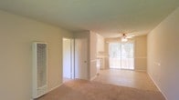 This single-floor residence offers a practical and comfortable layout with an open-concept living area that serves as the heart of the space. The living room features light-colored carpeting, neutral-toned walls, and a textured ceiling, creating a warm and inviting atmosphere. A ceiling fan is centrally positioned overhead, providing both air circulation and a subtle decorative element.

Natural light flows generously into the space through a large sliding glass door, which opens to an outdoor area and offers a pleasant view of surrounding greenery. Vertical blinds provide privacy and light control when needed.

The kitchen is conveniently situated adjacent to the living area, partially open to the main space via a pass-through counter. White cabinetry lines the upper portion of the kitchen, lending a clean and bright aesthetic. A closet door and a wall-mounted HVAC vent are also visible, highlighting the functional infrastructure integrated throughout the unit.

Overall, the space maintains a clean, straightforward design with consistent neutral tones and practical finishes, making it a well-organized and functional single-level living environment.