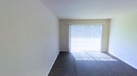 This single-floor space offers a clean and minimalist layout designed for comfortable living. The main living area immediately captures attention with its rich, dark hardwood flooring that contrasts elegantly against the light-toned walls and crisp white baseboards. A large sliding glass door, dressed with full-length vertical blinds, serves as the room's focal point, drawing in an abundance of natural light that brightens the entire space. The smooth, neutral-colored walls and ceiling provide a versatile backdrop well-suited for a variety of interior design styles. The open layout allows for flexible furniture arrangement, giving the space a sense of airiness and openness. The combination of the expansive glazed door and the warm tones of the flooring creates a welcoming ambiance throughout. Overall, the space presents a well-maintained, light-filled environment with timeless finishes that make it both functional and inviting.
