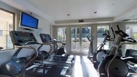 This fitness center is designed to provide a functional and motivating workout environment. The space is outfitted with a lineup of cardio equipment, including multiple treadmills and elliptical machines, arranged to maximize floor space and ease of movement. Natural light floods the room through large glass panel doors and wide windows that span the far wall, offering pleasant views of palm trees and an outdoor area beyond — creating an open, airy atmosphere that makes workouts more enjoyable. The light-toned flooring and white walls further enhance the bright, clean aesthetic of the space. A wall-mounted flat-screen television is positioned prominently for entertainment during exercise sessions. Recessed lighting in the ceiling supplements the abundant natural light, ensuring the space remains well-lit at any time of day. The glass doors provide direct access to the exterior, seamlessly connecting the indoor fitness area to the outdoor environment. Overall, the fitness center strikes a balance between practicality and comfort, offering a well-lit, thoughtfully arranged space suited for a variety of cardio-focused workouts.