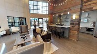This single-floor clubhouse blends modern sophistication with rustic warmth to create an inviting community gathering space. The main lounge area commands attention with soaring double-height ceilings, expansive steel-framed windows, and wide-plank hardwood flooring throughout. Plush seating arrangements, including tufted sofas and leather accent chairs, are thoughtfully positioned to encourage both relaxed conversation and social gatherings. A dramatic accent wall clad in reclaimed wood adds texture and character, anchoring the space with an organic, earthy feel. Adjacent to the lounge is a fully equipped wet bar and kitchen area, featuring gray cabinetry, crisp white quartz countertops, a herringbone tile backsplash, open floating shelves, stainless steel appliances, and a large French door refrigerator — all topped with a reclaimed wood ceiling detail that ties seamlessly into the overall design aesthetic. The recreation area offers a billiards table and media wall, framed by large steel-and-glass partitions that provide a visual connection to the adjoining fitness center, which is outfitted with exercise equipment and stability training tools. Generous natural light floods the interior through oversized windows and glass doors, which open to an outdoor patio area, further extending the space's social potential. The overall ambiance is refined yet approachable, balancing industrial and natural design elements throughout.