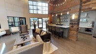 This single-floor clubhouse blends modern sophistication with rustic warmth to create an inviting community gathering space. The main lounge area commands attention with soaring double-height ceilings, expansive steel-framed windows, and wide-plank hardwood flooring throughout. Plush seating arrangements, including tufted sofas and leather accent chairs, are thoughtfully positioned to encourage both relaxed conversation and social gatherings. A dramatic accent wall clad in reclaimed wood adds texture and character, anchoring the space with an organic, earthy feel. Adjacent to the lounge is a fully equipped wet bar and kitchen area, featuring gray cabinetry, crisp white quartz countertops, a herringbone tile backsplash, open floating shelves, stainless steel appliances, and a large French door refrigerator — all topped with a reclaimed wood ceiling detail that ties seamlessly into the overall design aesthetic. The recreation area offers a billiards table and media wall, framed by large steel-and-glass partitions that provide a visual connection to the adjoining fitness center, which is outfitted with exercise equipment and stability training tools. Generous natural light floods the interior through oversized windows and glass doors, which open to an outdoor patio area, further extending the space's social potential. The overall ambiance is refined yet approachable, balancing industrial and natural design elements throughout.