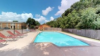 This outdoor amenity space centers around a generously sized swimming pool with clear, blue water and a classic geometric shape with angled corners. The pool is surrounded by a wide concrete deck that provides ample room for movement and relaxation. A row of red lounge chairs lines one side of the deck, offering residents a comfortable place to unwind and enjoy the sun. Adjacent to the pool area stands a warm-toned wooden cabana structure accented by a red umbrella and outdoor seating, providing a shaded gathering spot for socializing. The space is enclosed by a combination of wooden privacy fencing and a metal perimeter fence, creating a secure and intimate atmosphere. Lush, mature trees form a natural green backdrop that adds to the peaceful ambiance and offers partial shade throughout the day. The surrounding landscape is well-maintained, with open grassy areas visible beyond the fencing. The overall setting is bright and inviting, benefiting from abundant natural light and open sky views, making it an ideal outdoor retreat for warm-weather enjoyment.