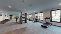Located at 585 North Ave in New Rochelle, NY, this well-appointed fitness center offers a comprehensive, single-floor wellness facility designed to support a wide range of workout routines. The space is divided into two distinct functional zones, seamlessly connected by glass double doors that enhance the open, airy feel of the environment.

The main workout area is generously proportioned and features patterned low-pile carpeting alongside large-format tile flooring. It is equipped with a diverse selection of cardio and strength machines, including multiple treadmills, an elliptical trainer, a rowing machine, a stationary bike, a multi-functional cable machine, a weight bench, and a cable pull-down station. Large dark-framed windows line the walls, flooding the space with natural light and offering views of the surrounding neighborhood. Full-length mirrors amplify the sense of space and allow for form-checking during workouts. Recessed ceiling lighting provides consistent illumination throughout.

Adjacent to the main gym is a dedicated yoga and stretching studio, outfitted with exercise mats and stability balls for flexibility and mindfulness training. Wall-mounted flat-screen TVs displaying calming imagery enhance the studio atmosphere. The clean white walls and neutral carpeting create a calm, focused environment ideal for low-impact exercise and recovery.
