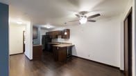 This single-floor space features an open-concept layout that seamlessly connects the kitchen and living area, creating a cohesive and functional environment. The kitchen is outfitted with rich, dark espresso-toned cabinetry that extends to the upper and lower sections, providing ample storage. Light-toned countertops offer a subtle contrast to the dark cabinetry, while a pendant light fixture above the peninsula adds focused illumination and a touch of style. A built-in wine cooler beneath the counter adds a practical and refined touch to the kitchen setup. Black appliances, including a refrigerator and dishwasher, complement the overall color palette. The flooring throughout is a dark hardwood-style material that anchors the space with warmth and continuity. A ceiling fan with an integrated light fixture is centrally positioned in the living area, contributing to both comfort and ambient lighting. The walls are finished in a clean, neutral white, enhancing the brightness of the space. Dark baseboards provide a polished frame that ties together the flooring and wall finishes, resulting in a cohesive and well-appointed interior.