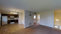 This single-floor residence offers a straightforward and functional layout designed for comfortable everyday living. The main living area is finished with wall-to-wall carpeting, providing a warm and inviting atmosphere, while neutral-toned walls keep the space feeling bright and open. A partial dividing wall separates the living area from the rest of the unit, adding a degree of structure without fully enclosing the space.

The kitchen is positioned adjacent to the living area and features dark wood cabinetry that contrasts with light countertops and white appliances, including a range. A decorative chandelier overhead adds a touch of character to this functional cooking space, and the flooring transitions to tile, making for easy maintenance. A ventilation duct and wall-mounted HVAC registers throughout indicate a centralized climate control system.

Beyond the partial wall, a hallway leads to additional rooms, with natural light visible through a window at the end of the corridor, suggesting the bedrooms benefit from outside light. The overall layout is practical and efficient, with clear sightlines connecting the key living and cooking spaces. This well-maintained unit presents a clean, move-in-ready environment suited for a variety of living arrangements.