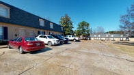 This fitness facility is set within a classic brick building distinguished by its dark mansard-style roof and traditional architectural detailing. A spacious paved parking lot fronts the property, providing convenient access for visitors. The surrounding area features mature trees that add a natural backdrop to the exterior.

Inside, the fitness space is thoughtfully organized into distinct workout zones, all unified by patterned, multi-tone blue carpeting. The main strength and conditioning area features soaring ceilings and is flooded with natural light through large windows fitted with white blinds, offering views of the surrounding landscape. This zone is equipped with a cable machine, power rack with bench, rowing machine, stationary bike, and a functional training rig complete with battle ropes, medicine balls, stability balls, and a full dumbbell rack.

A dedicated cardio area includes multiple treadmills positioned beneath a wall-mounted television, surrounded by windows on two sides. Large mirrors throughout the space enhance the open, airy ambiance. Recessed lighting and ceiling fans contribute to a comfortable training environment. An additional room with built-in shelving provides organized storage for accessories and supplies.