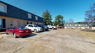 This fitness facility is set within a classic brick building distinguished by its dark mansard-style roof and traditional architectural detailing. A spacious paved parking lot fronts the property, providing convenient access for visitors. The surrounding area features mature trees that add a natural backdrop to the exterior.

Inside, the fitness space is thoughtfully organized into distinct workout zones, all unified by patterned, multi-tone blue carpeting. The main strength and conditioning area features soaring ceilings and is flooded with natural light through large windows fitted with white blinds, offering views of the surrounding landscape. This zone is equipped with a cable machine, power rack with bench, rowing machine, stationary bike, and a functional training rig complete with battle ropes, medicine balls, stability balls, and a full dumbbell rack.

A dedicated cardio area includes multiple treadmills positioned beneath a wall-mounted television, surrounded by windows on two sides. Large mirrors throughout the space enhance the open, airy ambiance. Recessed lighting and ceiling fans contribute to a comfortable training environment. An additional room with built-in shelving provides organized storage for accessories and supplies.