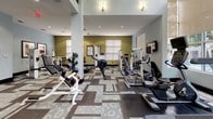 This fitness center offers a spacious, well-appointed environment designed to support a full range of workout routines. The space is characterized by impressively tall ceilings, with clerestory windows in the upper portion of one section allowing generous natural light to flood the interior, creating an open and energizing atmosphere. Additional windows fitted with horizontal blinds line the walls throughout, further enhancing the bright, airy feel.

The flooring features a distinctive patterned carpet in neutral tones of brown, gray, and cream, adding visual interest while providing a comfortable surface underfoot. Walls are painted in a two-tone scheme of soft sage green and light blue-gray, contributing to a calm, focused environment.

The cardio zone is well-stocked with elliptical trainers, treadmills, a rowing machine, and recumbent-style equipment from brands including Precor and Concept2. The strength training area includes selectorized cable machines, free weights, benches, and resistance equipment. Mirrors are strategically placed to expand the sense of space and assist with form. Recessed lighting supplements natural light throughout. A small built-in service counter and informational bulletin board near the entrance add functional convenience to the space.