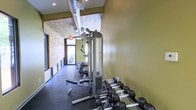 This fitness center offers a functional and well-appointed workout environment designed to support a full range of exercise routines. The space is characterized by a long, corridor-style layout that maximizes the available floor area while maintaining an open, airy feel.

Architecturally, the room stands out with its warm wood-planked ceiling, which adds a natural, rustic charm that contrasts beautifully with the smooth, olive-toned accent walls. Exposed industrial-style ductwork runs along the ceiling, lending a modern edge to the overall aesthetic. Large windows line one side of the space, allowing ample natural light to flood the interior and offering views of the greenery outside, creating an energizing atmosphere for workouts.

The fitness equipment is thoughtfully arranged throughout the space. A full dumbbell rack with a wide range of weights is prominently positioned for easy access, complemented by a multi-function cable machine suitable for a variety of strength training exercises. An adjustable workout bench and suspension training straps add further versatility. The rubber flooring throughout ensures safety and durability during high-impact activities. Recessed lighting supplements the natural light, keeping the space bright and inviting at all times.