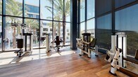 This expansive fitness center is designed with a contemporary aesthetic, offering a well-equipped and visually open environment for exercise. The space is defined by dramatic floor-to-ceiling glass walls that flood the interior with natural light and provide unobstructed views of palm trees and surrounding architecture, creating an energizing atmosphere throughout.

The flooring transitions between wood-look planks in the main exercise areas and rubber flooring in the dedicated weight training zone, providing both visual appeal and functional surfaces for varied workouts. Soaring double-height ceilings in portions of the facility enhance the sense of openness, while recessed lighting and an exposed industrial-style ceiling in the weight room add architectural contrast.

The strength training area is well-appointed with a full rack of dumbbells, benches, cable machines, and multi-use cable stations, complemented by large mirrors that visually expand the space. The cardio and selectorized machine zone runs along the glass perimeter, allowing users to work out with expansive outdoor views. Dark accent columns punctuate the space, adding structure and visual depth. Seating areas with upholstered benches provide rest zones, and signage indicates fitness policies, reflecting a professionally managed facility.