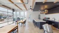 Located at 1904 Parable Way in Cary, NC, this single-floor community clubhouse blends modern farmhouse design with functional, open-concept living. The interior is defined by white shiplap walls, exposed wood ceiling beams, and patterned parquet-style flooring that flows throughout the space, creating a warm yet contemporary ambiance.

The kitchen is a standout feature, anchored by a large dark navy island with white quartz countertops and bar seating for several guests. The kitchen perimeter mirrors the island's cabinetry palette and is topped with matching counters, offering ample prep and storage space. Oversized woven pendant lights hang from a dramatic dark-painted ceiling, adding an artisan touch.

Multiple dining areas accommodate gatherings of varying sizes, including a large communal table near the garage-style glass doors. These floor-to-ceiling transparent doors open fully to the outdoor pool and patio area, flooding the interior with natural light and providing unobstructed views of the pool deck and surrounding greenery.

A cozy lounge area features neutral upholstered seating, a patterned area rug, a curated gallery wall, and a ceiling fan — ideal for relaxed conversation. Modern ceiling fans throughout the space ensure comfort year-round. The overall design is cohesive, airy, and inviting, making this clubhouse a well-appointed gathering destination for the community.