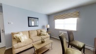 Located at 758 Christiana Rd in Newark, Delaware, this single-floor residence offers a calm and comfortable living environment designed with understated elegance. The living area sets a welcoming tone with soft blue-gray walls that create a relaxed, airy atmosphere. Neutral wall-to-wall carpeting runs throughout, providing warmth underfoot and a cohesive visual foundation for the space. A large window dressed with a decorative patterned valance allows generous natural light to fill the room, enhancing its open and pleasant feel. The room comfortably accommodates a full sofa and accent seating, complemented by a woven coffee table that adds a natural texture to the interior. A framed wall mirror with a dark trim is mounted on one wall, amplifying the sense of space and light. Baseboard heating and a wall-mounted HVAC unit ensure year-round comfort. The overall ambiance is modest yet inviting, with a neutral color palette and tasteful furnishings that create a cozy, livable atmosphere throughout this single-story home in Newark, Delaware.