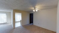 This single-floor residential space presents a straightforward, functional layout characterized by neutral tones and practical finishes throughout. The main living area features wall-to-wall beige carpeting that extends across the floor, contributing to a warm and understated aesthetic. A ceiling fan with an integrated light fixture is centrally positioned, providing both air circulation and ambient lighting to the room. The textured popcorn ceiling adds a layer of visual depth overhead.

The space connects to an adjoining room visible through an open doorway, where vertical blinds filter natural light, brightening that area considerably. This secondary room appears to feature a lighter flooring surface, offering a subtle contrast to the carpeted main area. A baseboard heater is visible along the wall, indicating a dedicated heating system. A dark-framed interior door leads to what appears to be a closet or additional room, adding to the overall functionality of the layout.

The walls are finished in a clean, neutral beige, keeping the space feeling open and versatile. Overall, the unit offers a practical and livable environment with essential features suited for comfortable, everyday residential use.