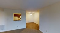 This single-floor residence offers a functional and straightforward layout designed for comfortable everyday living. The main living area features neutral-toned carpet underfoot and soft, light-colored walls that create a clean, open atmosphere throughout. A pass-through window connects the living space directly to the kitchen, providing an open sightline and a casual serving ledge. The kitchen is equipped with rich, warm-toned wood cabinetry, a white refrigerator, and a gas or electric range, all framed neatly within the pass-through opening. Adjacent to the main living area, a secondary room — accessible through an open doorway — features hardwood flooring and is equipped with a ceiling fan with an integrated light fixture, offering both ventilation and ambient lighting. The textured ceilings throughout the space add a subtle traditional character, while white baseboards provide a clean contrast against the flooring and walls. The overall design is neutral and move-in ready, with a practical flow between spaces that suits a variety of uses. The combination of carpet in the living zone and hardwood in the adjoining room offers both comfort and durability, making this a well-balanced and versatile single-story home.