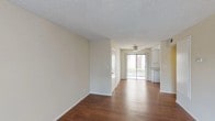 This single-floor residence offers a clean, functional layout with a clear separation between living, sleeping, and kitchen areas. The open living and dining space is finished with rich hardwood-style flooring that runs throughout the main areas, complemented by neutral cream-toned walls and a textured ceiling. A ceiling fan is centrally positioned, and a sliding glass door at the far end of the living area fills the space with natural light while providing direct access to an outdoor balcony or patio.

The kitchen is tucked to one side and features white cabinetry extending to the ceiling, granite-style countertops, a double-basin sink, a dishwasher, and a gas range, all set over matching hardwood-style flooring. A small window provides additional ventilation and light.

The bedroom is a well-proportioned, private room with soft carpeting, neutral walls, and a window fitted with vertical blinds that allow natural light while maintaining privacy.

The bathroom includes a wide vanity with granite-style countertops, white cabinetry, a mirrored medicine cabinet, and hardwood-style flooring. A separate wet area houses a full tub-shower combination, toilet, and a small window for natural ventilation. The overall space presents a simple, well-maintained interior.