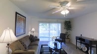 This single-story space presents a welcoming living area designed for both comfort and functionality. The room is anchored by a ceiling fan with an integrated light fixture, ensuring consistent airflow and illumination throughout. A large sliding glass door with vertical blinds serves as the room's focal point, allowing abundant natural light to fill the interior while providing direct views of a lush outdoor greenery. The light-toned walls and neutral carpeted flooring create a clean, cohesive backdrop that complements the room's furnishings. Seating includes a plush sofa adorned with patterned accent pillows and a coordinating armchair, arranged thoughtfully around a round glass-top coffee table. A geometric area rug grounds the seating arrangement and adds visual texture to the space. A tall decorative plant in the corner introduces a natural element, while a framed piece of wall art and a classic table lamp contribute warmth and personality. The textured ceiling adds a subtle architectural detail. Overall, the space offers a bright, functional, and comfortable environment with a relaxed, well-appointed ambiance.