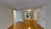 This single-floor space offers an open, flowing layout that seamlessly connects the living area to the kitchen. The most striking feature is the rich, warm hardwood flooring that runs throughout, polished to a high sheen that reflects light and adds a sense of warmth and depth to the interior.

The kitchen is positioned at the far end of the open space and is outfitted with stainless steel appliances, including a range, over-the-range microwave, and a full-size refrigerator. White cabinetry lines the walls, providing ample storage, while light-toned flooring in the kitchen area creates a subtle contrast with the hardwood in the main living space.

Neutral gray walls keep the interior feeling clean, bright, and versatile, lending themselves well to a variety of décor styles. Multiple doorways are visible, suggesting access to additional rooms or hallways, offering practical separation between living and private spaces. White trim and six-panel doors throughout contribute to a cohesive, classic aesthetic. The layout feels open and uncluttered, making efficient use of the available space on a single level.