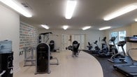 This fitness center offers a thoughtfully designed workout space that balances functionality with an energizing atmosphere. The room features a smooth, light-toned hard floor that transitions to a darker carpeted section in the cardio zone, providing a clear visual distinction between workout areas. Bright, recessed rectangular ceiling lights illuminate the entire space evenly, ensuring excellent visibility throughout.

The strength training area is anchored by a selection of dedicated resistance machines, including a prominent Instinct-branded unit and a multi-function cable station, catering to a full range of muscle groups. The cardio zone houses a lineup of upright and recumbent bikes alongside elliptical trainers and treadmills, offering diverse options for aerobic exercise.

A standout design element is the motivational mural covering one full wall, featuring bold typographic phrases in black and orange that inspire and energize those working out. Classic white French doors add an elegant architectural touch, bringing a sense of openness to the space. The combination of practical equipment layout, dynamic wall art, and clean, bright finishes creates a welcoming and motivating environment well-suited for a comprehensive fitness routine.