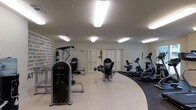 This fitness center offers a thoughtfully designed workout space that balances functionality with an energizing atmosphere. The room features a smooth, light-toned hard floor that transitions to a darker carpeted section in the cardio zone, providing a clear visual distinction between workout areas. Bright, recessed rectangular ceiling lights illuminate the entire space evenly, ensuring excellent visibility throughout.

The strength training area is anchored by a selection of dedicated resistance machines, including a prominent Instinct-branded unit and a multi-function cable station, catering to a full range of muscle groups. The cardio zone houses a lineup of upright and recumbent bikes alongside elliptical trainers and treadmills, offering diverse options for aerobic exercise.

A standout design element is the motivational mural covering one full wall, featuring bold typographic phrases in black and orange that inspire and energize those working out. Classic white French doors add an elegant architectural touch, bringing a sense of openness to the space. The combination of practical equipment layout, dynamic wall art, and clean, bright finishes creates a welcoming and motivating environment well-suited for a comprehensive fitness routine.
