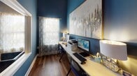 This single-floor amenity space is thoughtfully designed to serve as a multifunctional community hub. The interior is unified by a striking deep blue color palette applied to the walls, creating a cohesive and modern aesthetic throughout. Warm hardwood flooring runs continuously across the space, complementing the bold wall tones and adding a welcoming sense of warmth.

The business center is a dedicated workspace equipped with multiple computer workstations along a built-in countertop desk. Decorative table lamps and a large abstract artwork with gold accents lend an upscale feel to the area. Natural light filters in through a window dressed with gold-patterned sheer curtains, brightening the room. A printer and posted rules of use signage indicate this is a shared, fully functional workspace.

Adjacent areas are visible through large glass pass-through openings framed in white trim, connecting the business center to a lounge and seating area outfitted with dark upholstered chairs, a bar-height table with stools, and a wall-mounted television. Beyond, a fitness area with exercise equipment is accessible through a glass-paneled door, making the overall space a comprehensive amenity suite designed for both productivity and leisure.