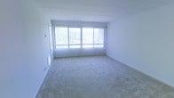 This single-floor space presents a clean, minimalist layout centered around a generously sized main living area. The room is finished with wall-to-wall carpet in a neutral beige tone, providing a soft, understated foundation that complements the overall bright aesthetic. White walls and a white ceiling contribute to a crisp, open atmosphere throughout.

A prominent feature of the space is the expansive bank of windows spanning much of one wall, fitted with vertical blinds that allow for adjustable natural light. The windows invite an abundance of daylight into the room, enhancing the sense of openness and making the space feel larger than its footprint. A glimpse of greenery through the glass suggests outdoor foliage nearby, adding a subtle connection to the outdoors.

The room's layout is straightforward and versatile, lending itself well to a variety of furniture arrangements. A doorway is visible along one wall, indicating connectivity to adjacent areas of the unit. The overall condition of the space appears well-maintained and move-in ready, offering a neutral canvas suited to a range of interior design preferences.