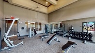 This fitness center in Conroe, TX is designed to support a full range of workout routines, divided into two distinct areas: a weights zone and a dedicated yoga studio. The weights area impresses with soaring ceilings and an open, airy atmosphere enhanced by large windows that flood the space with natural light and offer views of the surrounding greenery. Floor-to-ceiling mirrors line the walls, visually expanding the room and allowing users to monitor their form. The space is outfitted with a comprehensive selection of strength training equipment, including Hoist cable machines, TRX suspension trainers, a full dumbbell rack ranging across multiple weights, adjustable benches, and a variety of selectorized machines for targeted muscle training. Durable carpet tile flooring provides comfort and stability underfoot throughout the workout area. The adjacent yoga studio offers a serene, open-plan environment bathed in natural light from tall windows. A striking, large-scale yoga-themed wall mural serves as the room's focal point, complemented by a mounted display screen for guided sessions. The high ceilings in both areas contribute to an expansive, motivating atmosphere, making this fitness center a versatile and inviting facility for strength training and mindful movement alike.
