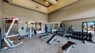 This fitness center in Conroe, TX is designed to support a full range of workout routines, divided into two distinct areas: a weights zone and a dedicated yoga studio. The weights area impresses with soaring ceilings and an open, airy atmosphere enhanced by large windows that flood the space with natural light and offer views of the surrounding greenery. Floor-to-ceiling mirrors line the walls, visually expanding the room and allowing users to monitor their form. The space is outfitted with a comprehensive selection of strength training equipment, including Hoist cable machines, TRX suspension trainers, a full dumbbell rack ranging across multiple weights, adjustable benches, and a variety of selectorized machines for targeted muscle training. Durable carpet tile flooring provides comfort and stability underfoot throughout the workout area. The adjacent yoga studio offers a serene, open-plan environment bathed in natural light from tall windows. A striking, large-scale yoga-themed wall mural serves as the room's focal point, complemented by a mounted display screen for guided sessions. The high ceilings in both areas contribute to an expansive, motivating atmosphere, making this fitness center a versatile and inviting facility for strength training and mindful movement alike.