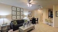This single-floor residence showcases a thoughtfully designed open-concept layout that seamlessly connects the living and dining areas, creating a welcoming and functional living environment. The living area is anchored by plush carpeting in warm neutral tones, complemented by a ceiling fan with integrated lighting that adds both comfort and ambiance. A gallery-style arrangement of framed botanical prints adorns one wall, lending the space a curated, sophisticated feel. The centerpiece of the room is a dark upholstered sofa paired with a contemporary coffee table, styled with decorative glass vessels and greenery for an elevated touch. Track lighting runs along the hallway ceiling, providing directional illumination that enhances the flow between spaces. Adjacent to the living area, a compact dining space features a dark round table and matching chairs, ideal for intimate gatherings. A decorative wall piece adds visual interest, while neutral-painted walls throughout maintain a clean, cohesive aesthetic. The hallway leads toward additional rooms, hinting at a well-organized floor plan. Crown molding along the ceiling adds a refined architectural detail, tying the overall design together with a polished finish. The space strikes a balance between comfort and style.