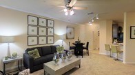 This single-floor residence showcases a thoughtfully designed open-concept layout that seamlessly connects the living and dining areas, creating a welcoming and functional living environment. The living area is anchored by plush carpeting in warm neutral tones, complemented by a ceiling fan with integrated lighting that adds both comfort and ambiance. A gallery-style arrangement of framed botanical prints adorns one wall, lending the space a curated, sophisticated feel. The centerpiece of the room is a dark upholstered sofa paired with a contemporary coffee table, styled with decorative glass vessels and greenery for an elevated touch. Track lighting runs along the hallway ceiling, providing directional illumination that enhances the flow between spaces. Adjacent to the living area, a compact dining space features a dark round table and matching chairs, ideal for intimate gatherings. A decorative wall piece adds visual interest, while neutral-painted walls throughout maintain a clean, cohesive aesthetic. The hallway leads toward additional rooms, hinting at a well-organized floor plan. Crown molding along the ceiling adds a refined architectural detail, tying the overall design together with a polished finish. The space strikes a balance between comfort and style.