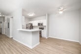 This single-floor residence offers a clean, modern design with a cohesive neutral color palette and light-toned luxury vinyl plank flooring throughout the main living areas. The open-concept layout connects the living room and kitchen seamlessly, creating a bright and functional shared space. The living room features a ceiling fan and a sliding glass door fitted with vertical blinds, allowing natural light to fill the room while providing access to an outdoor area.

The kitchen is a standout feature, designed in a galley style with crisp white shaker cabinetry, light quartz countertops, and a full suite of stainless steel appliances including a refrigerator, electric range, over-the-range microwave, and dishwasher. A peninsula with a double sink adds additional prep space and casual seating potential, reinforcing the open flow between the kitchen and living areas.

The bedrooms are carpeted for comfort and include windows with blinds that bring in natural light. Generous closet space with wire shelving is provided in the bedrooms. The bathroom features a white vanity with quartz countertops, a single sink, a large mirror, and chrome fixtures, all set against light flooring and neutral walls. A dedicated hallway connects all rooms, contributing to an efficient and practical floor plan throughout.