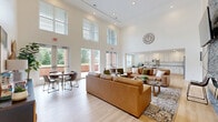 Located at 7300 Ashlake Pkwy in Chesterfield, VA, this single-floor open-concept space is designed to impress with its dramatic double-height ceilings that amplify the sense of openness throughout. The main gathering area serves as the centerpiece, anchored by a floor-to-ceiling stacked stone accent wall housing a fireplace and wall-mounted television — a bold architectural focal point that adds texture and warmth to the bright, neutral interior.

Light hardwood flooring runs throughout, complementing the crisp white walls and recessed lighting overhead. Oversized windows along the perimeter flood the space with natural light and offer pleasant views of the surrounding greenery, creating an inviting, airy ambiance. Multiple seating arrangements — including plush leather sofas and accent chairs — provide flexible areas for lounging and conversation, while a round dining table offers a more intimate setting near the glass entry doors.

The fully equipped kitchen is a standout feature, outfitted with white shaker cabinetry, granite countertops, stainless steel appliances including a range, refrigerator, and dishwasher, and a large island with bar seating — ideal for both casual dining and food preparation. Additional rooms accessible via glass-panel doors suggest supplementary utility or meeting spaces. The overall design strikes a balance between modern sophistication and welcoming comfort.