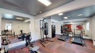 Located at 929 Nord Avenue in Chico, California, this fitness facility offers a well-organized and fully equipped exercise space on a single floor. The exercise room is the heart of the facility, featuring a dark, textured ceiling that contrasts with clean white walls accented by bold red detailing — lending the space an energetic, motivating atmosphere. Multicolored modular carpet tiles cover the floor throughout, providing both durability and cushioning underfoot. The room is illuminated by large flush skylights and recessed lighting, ensuring bright, even coverage across the workout area. Equipment includes multiple treadmills, a rowing machine, a stationary bike, and a heavy punching bag on a freestanding stand, catering to a variety of cardio and strength training routines. A large wall mirror at one end amplifies the sense of space and supports exercise form. A small desk area with a window view to the outdoors provides a practical check-in or administrative spot. The facility also includes a compact, cleanly finished bathroom with a wall-mounted sink, toilet, and dark-toned flooring — functional and neatly maintained. Natural light filters in through several windows, connecting the interior to the surrounding greenery outside.