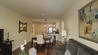 This single-floor residence offers a thoughtfully designed open-concept layout that seamlessly connects the living, dining, and kitchen areas into one cohesive and inviting space. The interior is finished in warm neutral tones throughout, creating a calm and sophisticated ambiance that feels both comfortable and refined.

The living area is anchored by a plush upholstered sofa and complemented by an elegant table lamp, setting a welcoming tone. It flows naturally into the dining space, which features a dark wood dining table surrounded by cushioned upholstered chairs — ideal for everyday meals or entertaining guests. Dark hardwood flooring runs throughout, adding warmth and visual continuity across the open floor plan.

The kitchen is situated at the rear of the space, partially open to the dining area via a raised breakfast bar that provides both a casual seating option and a natural visual divider. The kitchen is equipped with dark appliances, ample cabinetry in a light wood finish, and is illuminated by a series of stylish pendant lights that hang at varying heights, adding a decorative focal point overhead. Recessed lighting in the ceiling provides even, ambient illumination throughout the space.

A decorative mirror on the living room wall adds depth and light, while architectural details such as clean lines and structured room transitions contribute to the overall polished character of this well-designed residence.