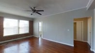 This single-story space showcases a blend of classic architectural details and functional design. The main living area is defined by rich, dark hardwood floors that extend throughout, lending warmth and a timeless aesthetic to the space. Soft gray walls provide a neutral backdrop that complements the natural textures found throughout the room.

A standout feature is the exposed brick wall, which adds a distinctive, rustic character to the interior. Natural light filters in through horizontal-slatted blinds on the windows, brightening the space while offering privacy. A ceiling fan with a light fixture is centrally mounted on the textured ceiling, which features classic crown molding along the perimeter, adding a refined architectural touch.

An open doorway leads to an adjacent area, where glimpses of a classic black-and-white checkered tile floor are visible, suggesting a transitional space such as an entryway or hallway. White trim and baseboards frame the walls cleanly throughout, creating a crisp contrast against the gray tones. The overall ambiance of the space is understated and inviting, with a balance of vintage charm and clean, neutral finishes.