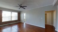 This single-story space showcases a blend of classic architectural details and functional design. The main living area is defined by rich, dark hardwood floors that extend throughout, lending warmth and a timeless aesthetic to the space. Soft gray walls provide a neutral backdrop that complements the natural textures found throughout the room.

A standout feature is the exposed brick wall, which adds a distinctive, rustic character to the interior. Natural light filters in through horizontal-slatted blinds on the windows, brightening the space while offering privacy. A ceiling fan with a light fixture is centrally mounted on the textured ceiling, which features classic crown molding along the perimeter, adding a refined architectural touch.

An open doorway leads to an adjacent area, where glimpses of a classic black-and-white checkered tile floor are visible, suggesting a transitional space such as an entryway or hallway. White trim and baseboards frame the walls cleanly throughout, creating a crisp contrast against the gray tones. The overall ambiance of the space is understated and inviting, with a balance of vintage charm and clean, neutral finishes.