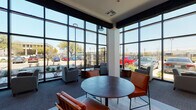 This contemporary single-floor space is defined by its striking architectural design, most notably the expansive floor-to-ceiling window walls that wrap around the lounge area, flooding the interior with natural light and offering unobstructed views of the surrounding landscaped exterior and streetscape. The soaring ceiling height creates an open, airy atmosphere that feels both welcoming and sophisticated.

The main lounge area features a thoughtfully curated mix of seating arrangements, including plush gray upholstered chairs, sleek cognac leather accent chairs with black metal frames, and round wooden side tables. A combination of light hardwood-style flooring and soft carpeted areas defines distinct zones within the space, adding texture and visual interest underfoot.

Decorative elements are carefully selected to complement the modern aesthetic. Bold abstract wall art in teal, olive, and navy tones adds color and personality, while striking starburst wall sculptures serve as eye-catching focal points. Exposed dark wood ceiling beams and tall cylindrical pendant lighting contribute warmth and contrast to the otherwise crisp white walls and open framework.

Glass-paneled interior doors with black frames provide access to adjacent areas while maintaining visual connectivity. Potted greenery and landscaped plantings both inside and outside the window line bring a natural element into the design, completing the polished, contemporary ambiance of this well-appointed space.