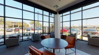 This contemporary single-floor space is defined by its striking architectural design, most notably the expansive floor-to-ceiling window walls that wrap around the lounge area, flooding the interior with natural light and offering unobstructed views of the surrounding landscaped exterior and streetscape. The soaring ceiling height creates an open, airy atmosphere that feels both welcoming and sophisticated.

The main lounge area features a thoughtfully curated mix of seating arrangements, including plush gray upholstered chairs, sleek cognac leather accent chairs with black metal frames, and round wooden side tables. A combination of light hardwood-style flooring and soft carpeted areas defines distinct zones within the space, adding texture and visual interest underfoot.

Decorative elements are carefully selected to complement the modern aesthetic. Bold abstract wall art in teal, olive, and navy tones adds color and personality, while striking starburst wall sculptures serve as eye-catching focal points. Exposed dark wood ceiling beams and tall cylindrical pendant lighting contribute warmth and contrast to the otherwise crisp white walls and open framework.

Glass-paneled interior doors with black frames provide access to adjacent areas while maintaining visual connectivity. Potted greenery and landscaped plantings both inside and outside the window line bring a natural element into the design, completing the polished, contemporary ambiance of this well-appointed space.
