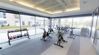 This contemporary fitness center offers a well-appointed exercise space designed to inspire and motivate. The cycling studio takes center stage, featuring a fleet of professional-grade stationary bikes with bold yellow accents, arranged to provide ample space between each station. A dedicated free weights area includes a neatly organized kettlebell rack, catering to a variety of workout styles.

The space is wrapped in expansive floor-to-ceiling windows on multiple sides, flooding the interior with natural light and offering open views of the surrounding outdoor terrace and landscaped grounds. Roller shades provide optional light control while maintaining the bright, airy atmosphere.

The ceiling design is a standout architectural feature, showcasing an exposed concrete finish with warm indirect cove lighting that frames the perimeter, lending the space an upscale, industrial-modern aesthetic. The flooring transitions between smooth and textured rubber surfaces, delineating functional zones while providing comfort and safety underfoot.

Adjacent to the cycling area, a lounge zone with comfortable seating offers a space to cool down or socialize. The overall ambiance is sleek, energetic, and professional, making this fitness center a premium amenity suited for a high-end residential or commercial environment.