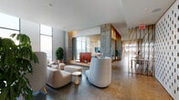 This thoughtfully designed single-floor space blends industrial and contemporary elements to create a refined, multifunctional environment. Exposed concrete columns and polished concrete floors establish a raw yet refined architectural foundation throughout, while soaring floor-to-ceiling windows flood the lounge area with abundant natural light and offer sweeping panoramic city views.

The main lounge is anchored by a generous curved sectional sofa and sculptural high-back chairs arranged on a circular area rug, creating an inviting yet sophisticated seating environment. A glass-enclosed private room, lined with warm wood paneling and featuring its own television and dining area, provides a more intimate setting separated from the open floor plan.

The bar and dining area centers on a large, white-topped island with bar stool seating, complemented by warm wood cabinetry, a multi-tap draft station, and a dramatic large-scale artwork mounted above a continuous backlit countertop. Globe pendant lights illuminate the space overhead.

A distinctive perforated white panel wall serves as both a design feature and spatial divider, adding graphic texture across the room. The overall ambiance is contemporary and curated, balancing communal openness with defined zones for socializing, dining, and private gatherings.