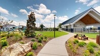 Located at 9035 Minnow Mews in Rowlett, TX, this community fieldhouse spans 2,560 square feet across three floors — 1,900 sq ft on the ground floor, 590 sq ft on the upper level, and an 80 sq ft mezzanine level. The building's modern farmhouse exterior features board-and-batten siding, a metal roof, and large glass entry doors framed by welcoming stepped concrete approach. Inside, the fitness center commands attention with soaring vaulted ceilings clad in warm wood paneling, linear LED lighting, floor-to-ceiling mirrors, and a full array of cardio and strength equipment. A cozy lounge nook offers a cushioned seating area accented with local Texas-themed artwork. The expansive covered outdoor patio showcases a striking wood-paneled vaulted ceiling with ceiling fans, comfortable lounge seating, outdoor dining, and a built-in grilling station — ideal for community gatherings. An adjacent covered basketball court extends the recreational amenities. The surrounding neighborhood features meticulously landscaped streetscapes with native plantings, mulched garden beds, bike racks, decorative street lighting, and a tranquil community pond, all connected by wide concrete walking paths.