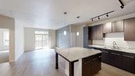 This well-designed single-floor residence offers a seamless blend of modern finishes and functional layout. The open-concept kitchen and living area serves as the heart of the space, anchored by a striking granite-topped island with dark wood support columns and ample cabinetry storage beneath. Rich dark espresso cabinetry lines the kitchen walls, complemented by light granite countertops, stainless steel appliances including a refrigerator, microwave, and range, and a clean tile backsplash. Track lighting overhead and elegant pendant lights above the island create a warm, layered ambiance. Light wood-look flooring extends throughout the main living area, lending a bright and cohesive feel. The living room opens directly to a private balcony through sliding glass doors, inviting natural light and outdoor views into the space. The bedrooms are finished with plush neutral carpeting and large windows fitted with roller shades that provide both light and privacy. The bathroom features warm stone-toned tile flooring, a walk-in shower with chrome fixtures, a dark wood vanity with a white countertop, and clean white walls that maintain a crisp, contemporary aesthetic throughout.