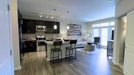 This well-designed single-floor space showcases a seamless open-concept layout that connects the kitchen and living area into one cohesive and functional environment. The kitchen is appointed with dark espresso cabinetry, stainless steel appliances including a range, microwave, and refrigerator, complemented by a light-toned countertop and a subtle tile backsplash. A central kitchen island with bar seating provides an ideal spot for casual dining or entertaining. Pendant lighting hangs above the island, adding a warm, modern touch, while track lighting overhead ensures the kitchen is well-illuminated throughout. The living area flows naturally from the kitchen, featuring wide-plank hardwood-style flooring that extends across the entire space, creating a unified and polished aesthetic. A large window along the far wall allows generous natural light to fill the room, enhancing the open and airy atmosphere. The space is accented with contemporary décor, including a mixed-media wall art piece that adds a pop of color and character. The overall ambiance is modern and inviting, balancing stylish design with practical, everyday functionality.